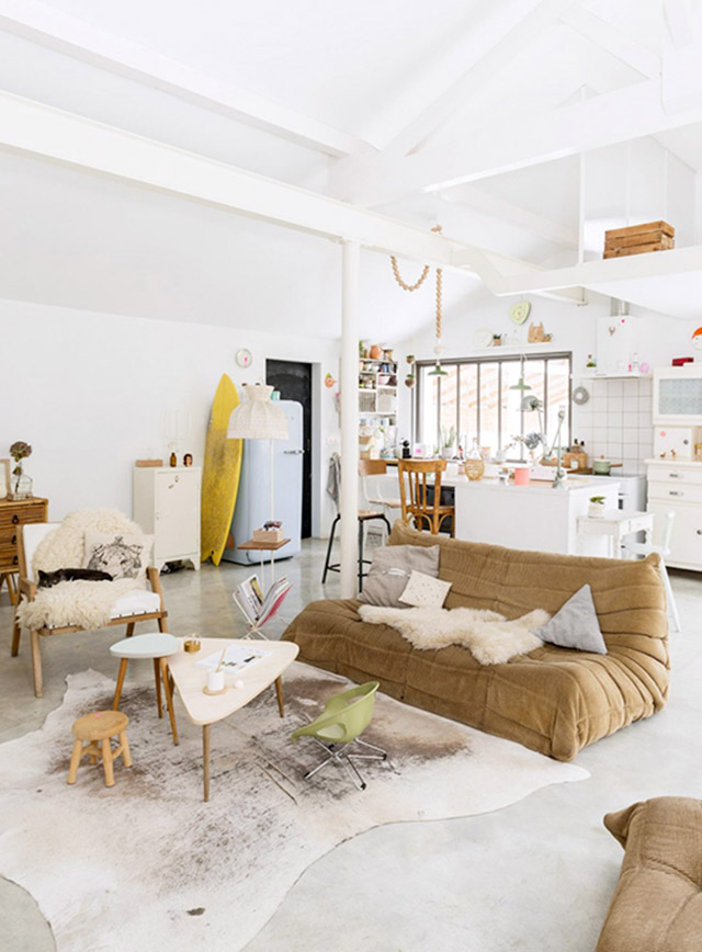 Vista a un gran living y estar con la cocina americana al fondo. Decoración en blancos y ocres, con muebles de líneas simples en la cocina, living con sofás de tela sin patas en color amarillo mostaza y mesas de centro triangulares con patas de madera.