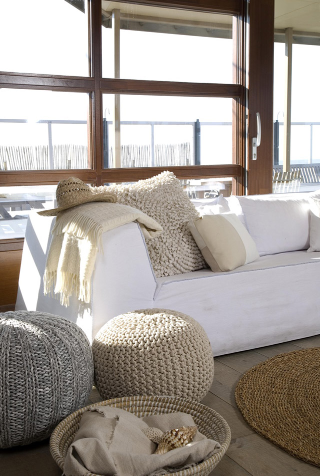 Sala de estar de casa playera con ventanales de marco de madera. Sofá de tela blanca con cojines y manta en tonos blancos y colores crema o arena. Poufs de lana tejidos en colores cremas, canastos y alfombras de fibra natural.