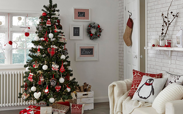 Muro decorado con un par de cuadros con motivos navideños, una corona navideña, pino con regalos y repisa con adornos rojos y blancos.