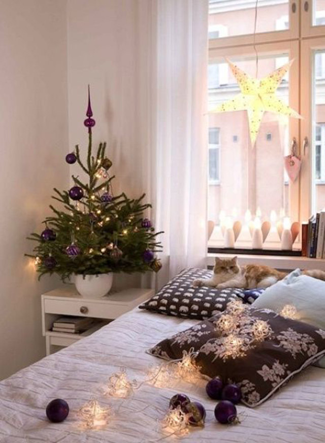 Dormitorio decorado en estilo navideño. Junto a la ventana que da al respaldo de la cama, porta velas en forma de ángeles y una lámpara de estrella colgando del marco de la ventana. Sobre el velador un pequeño pino con adornos violetas y luces y, en la cama guirnalda de luces en forma de farolitos y esferas decorativas color violeta.