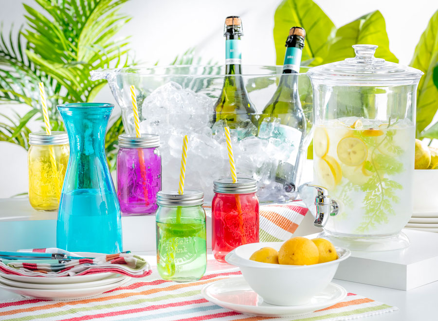 Mesa con vasos y jarras de colores con una gran hielera transparente y un recipiente para jugo de vidrio lleno de agua y limones