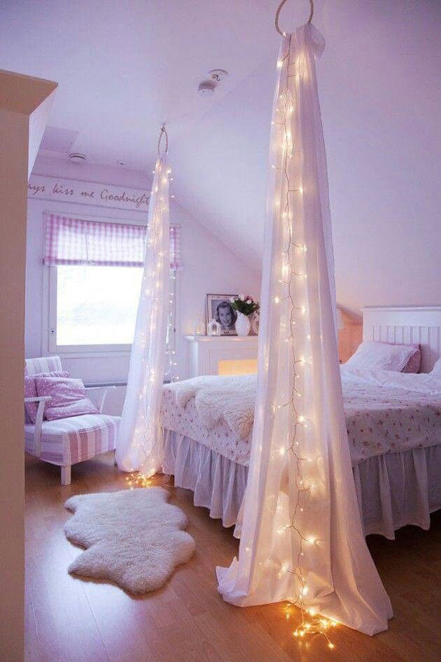 Dormitorio estilo romántico en color lila, poltrona con tela listada en lila y blanco, bajada de cama de piel, a la altura de los pies de la cama y desde el techo, cualgan unas cortinas blancas con guirnaldas de luces.