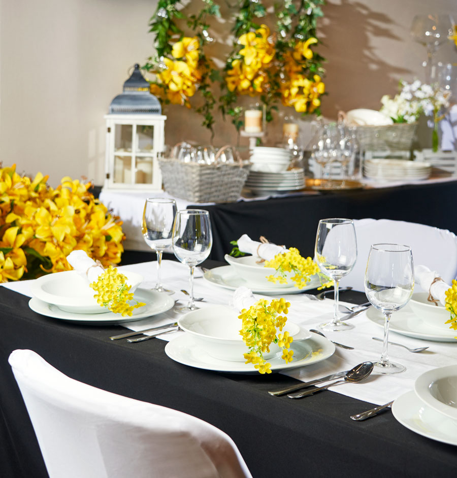 Mesa de comedor con mantel negro, loza blanca adornada con flores amarillas. Copas transparentes y vajilla plateada