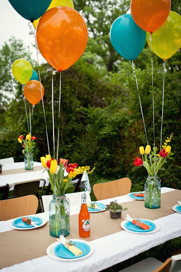 Centros de mesa de frascos de vidrio celeste. Cada centro de mesa tiene tulipanes rojos y amarillos y tres globos con helio de colores anaranjado, celeste y amarillo.