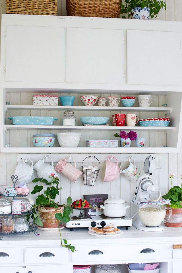 Mueble de cocina blanco con puertas y además repisas a la vista. En las repisas se ve vajilla de colores celeste, rojo y blanco con diseños de puntitos y de flores. Sobre el mesón un perchero con jarrones colgados de colores rosado y blanco.