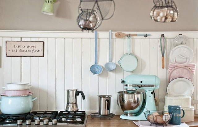 Vista frontal de cocina con los quemadores a un lado y al otro el mesón con una batidora metálica de color turquesa, cajas metálicas rosadas, canasta con huevos, muro con ganchitos con utensilios de latón esmaltado en colores turquesa y celeste.