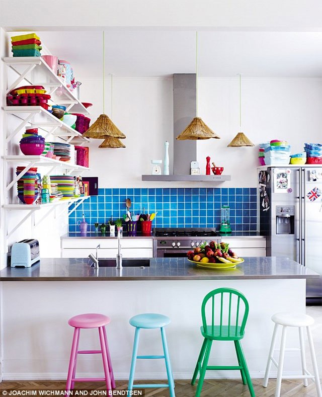 Vista general de cocina estilo americana con muros blancos y en la zona de los quemadores y la campana cerámica azul eléctrico. El mesón donde está el lavaplatos tiene alrededor pisos de colores rosado, celeste y blanco y una silla verde. Repisas con vajilla y utensilios de colores rojos, verdes, violetas y amarillos. Lámparas colgantes con pantallas de fibra natural.