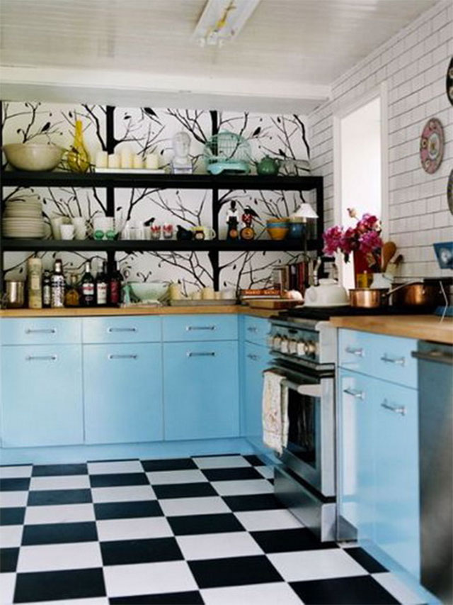 Cocina con muros de ladrillo pintado blanco, muebles de cocina color celeste, encimera de madera, piso de baldosas blanco y negro. Uno de los muros con un fotomural de árboles en blanco y negro, repisa negra con utensilios en blanco, celeste y amarillos.