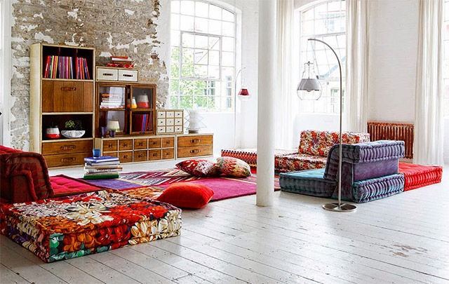 Vista general de un living en un loft con ventanales en una esquina y muebles multicolores tipo sofás cama estampados con motivos florales en rojo y blanco, otros de color azul/violeta y rojos.