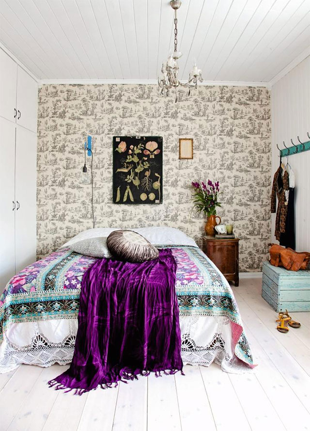 Dormitorio muros laterales y techo blancos, muro de fondo en papel mural motivo ilustración de árboles en blanco y negro. Cama con faldón blanco con bordes broderie, cobertor diseño floral en colores celeste, negro, violetas y blanco, manta púrpura, cojín gris y cojín café y blanco.