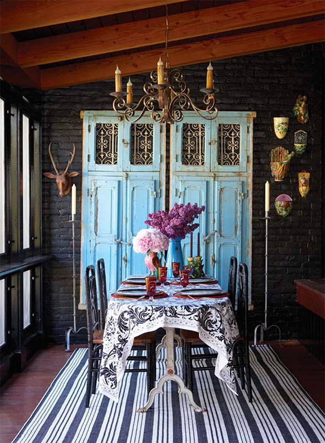 Comedor con mesa ovalada, mantel blanco con diseño floral en negro, sillas negras con respaldos torneados, asientos de madera natural, al final de la mesa un florero esmaltado color calipso. En la pared de ladrillos pintados negro un par de puertas talladas color celeste, al costado  una cabeza de madera de un ciervo. Cuelga sobre la mesa una lámpara tipo candelabro. Alfombra listada azul con blanco.