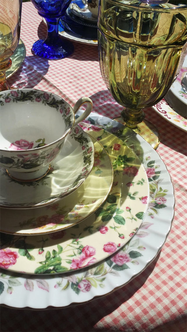 Vajilla de porcelana floreada, copas de vidrio verde sobre mantel cuadrillé rojo con blanco.