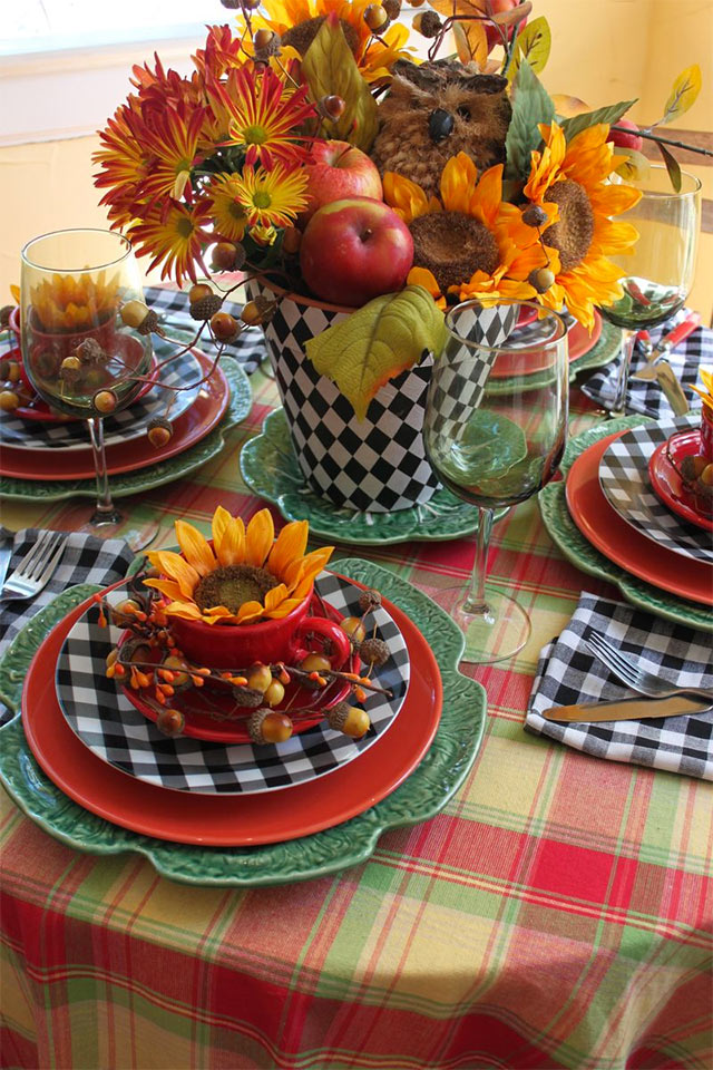 Mesa redonda con mantel escocés rojo, verde y amarillo. Vajilla con plato grande verde en forma de flor, plato mediano rojo, plato pequeño a cuadros negro con blanco y finalmente taza y plato rojo decorado con un girasol. Copas de vidrio en tono verde