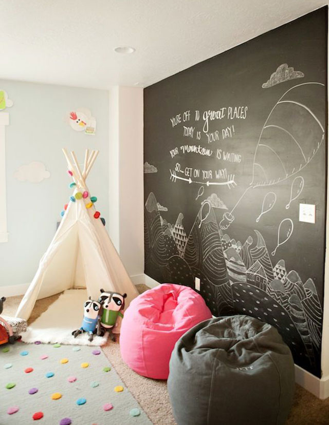 Habitación infantil con alfombra gris y círculos de colores, dos poufs o peras, una rosada y la otra gris, una carpita tipo india de lona blanca y guirnaldas de bolitas de colores. Un muro completo negro pizarrón donde se puede dibujar.