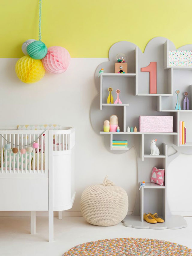 Dormitorio infantil con muro blanco y la cuarta parte hacia el cielo pintada amarillo, cuelgan unos pompones grandes de papel en colores rosado, blanco, verde menta y amarillo. Estante/repisa en forma de árbol pintado gris pálido con accesorios y juguetes en colores similares a los pompones.