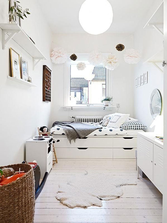 Dormitorio niña con cama al fondo junto a la ventana. Cama casi al ancho de la habitación, con cajones en la base. Decoración con predominio del blanco en muros y muebles. Textil cama y cubierta de un velador de lunares negros. Sobre la cama y desde el techo cuelgan pompones de papel en color rosa pálido.