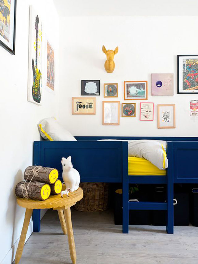 Cama de madera tipo cajón de color azul intenso con una pequeña escalera, sábanas amarillas contrastando con el azul. Decoración mural de cuadros con ilustraciones. Mesita lateral en color mostaza y encima apilados unos troncos y animalitos de juguete.