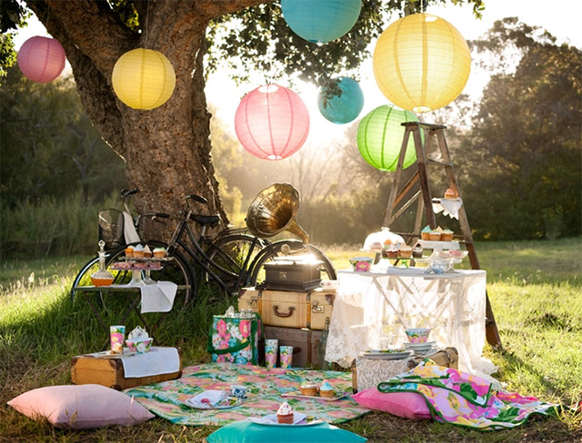 Pic nic junto a un gran árbol del que cuelgan lámparas de papel de colores, un par de bicicletas apoyadas en el árbol. Una mesita plegable con cupcakes junto a una torre de maletas y encima un gramófono. En el piso una manta floreada rodeada de cojines de colores rosado, calipso y fucsia.