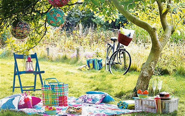 Picnic durante un día soleado. SObre el pasto una manta floreada en colores fuscia, rosado y blanco tipo patchwork, cojines en los tonos de la manta, canasta de picnic de mimbre, una silla plegable azul con un farol fucsia, todo esto junto a un árbol del que cuelgan lámparas circulares con diseños floreados.