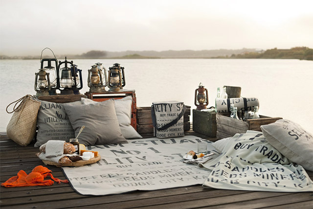 Picnic en un muelle a la orilla del lago. Cajas de madera cercando la zona, sobre estas cajas, faroles o linternas de parafina. Manta de tela gris con letras estampadas, cojines a tono.