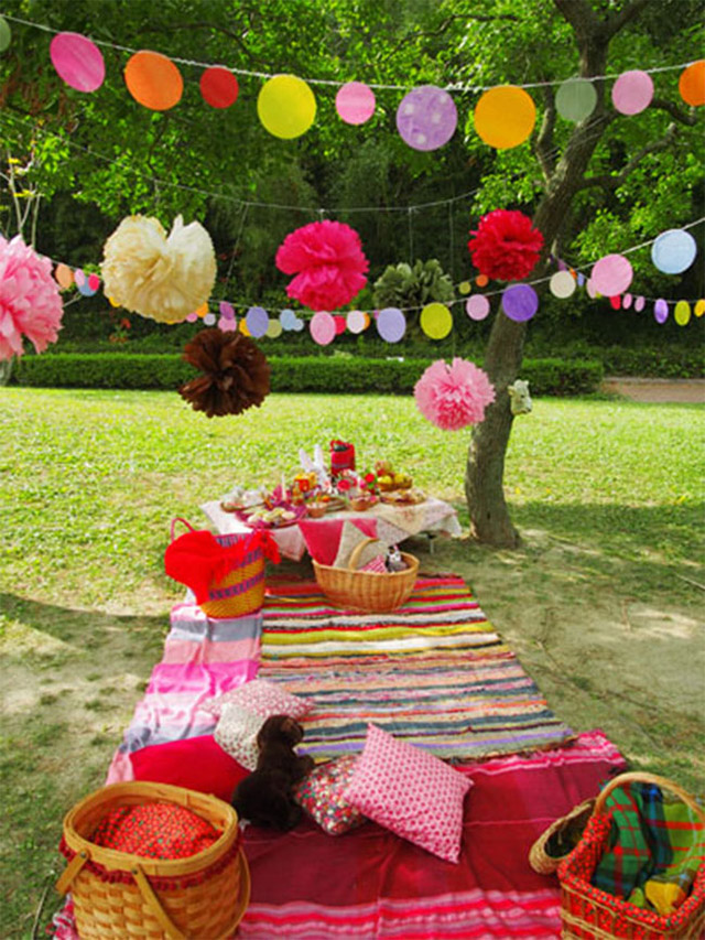 Fiesta de color estilo picnic en el parque. Varias mantas de colores fusia, rojo, amarillo, dispuestas a lo largo como si fuera una gran mesa. Al final se ve una mesa baja con la comida. Cojines a tono y canastos de fibras naturales con mantitas para el abrigo. Decoración aérea multicolor de guirnaldas de papel.