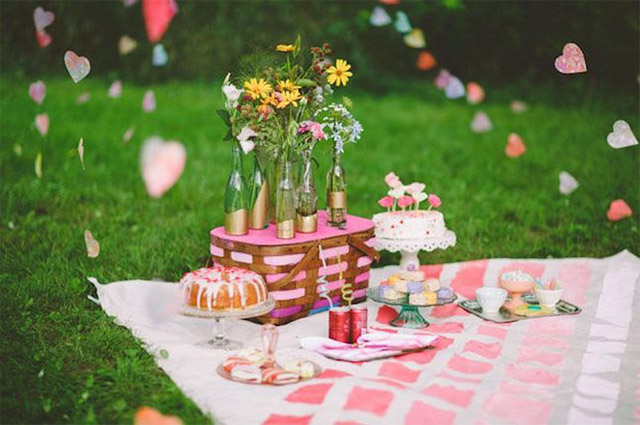 Canasta de picnic tejida de madera y tela, sobre la tapa de la canasta botellas con flores, todo esto sobre una manta en tonos rosados. Pasteles y tortitas, decoración aérea con guirnaldas de corazones de papel.