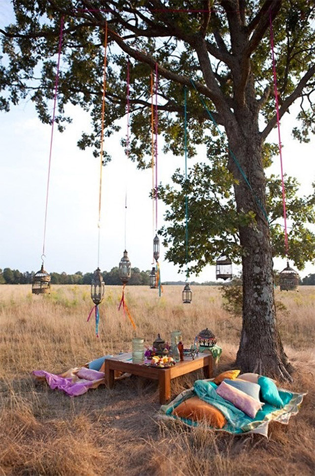 Mesita baja de madera, mantas de colores a ambos lados de la mesita, ésta decorada con jarrones de vidrio y canasto con flores. De un árbol que se encuentra junto cuelgan unos faroles con cintas de colores.