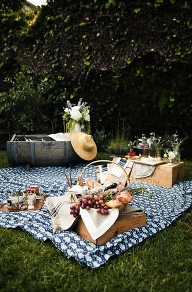 Picnic en tonos verde y azul. El verde lo dan los árboles, arbustos y césped. Sobre el césped una manta estampada en color azul y blanco. Canasta con frutas, la mitad de un barril con refrescos y sobre una caja botellas con flores