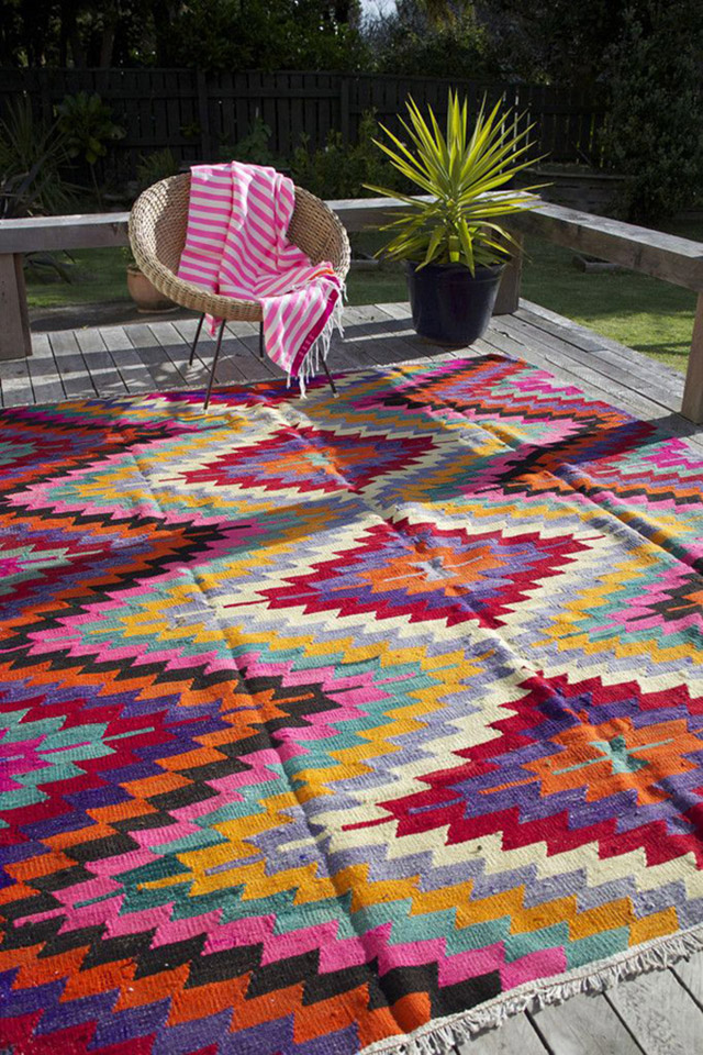 Amplia terraza de piso de madera saliendo a un jardín. Silla de mimbre y patas metálicas y alfombra multucolor con diseño geométrico.