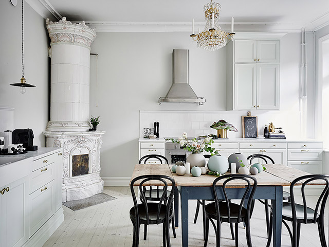 Cocina con comedor incorporado. Muebles de la cocina en blanco, chimenea estilo rococó en una esquina, lámpara de lágrimas en el techo. La mesa de comedor de madera con la base pintada en azul y la superficie al natural más sillas negras.