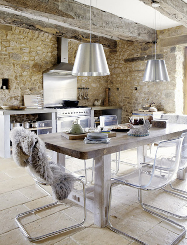 Cocina con muros de piedra y vigas a la vista, mezclado con muebles de cocina en blanco, madera natural y accesorios cromados. El comedor se encuentra dentro de la cocina con una mesa de madera en color natural y sillas de estructura metálica y respaldo y asiento transparentes. Sobre la mesa dos lámparas de pantalla plateada estilo industrial.