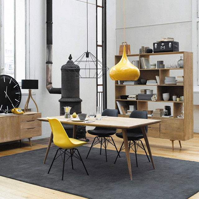 Comedor estilo industrial con mesa de comedor de madera color natural y sillas tipo Eames, dos sillas negras y una amarilla. Alfombra color grafito, lámpara de pantalla amarilla sobre la mesa y muebles estilo deco alrededor.