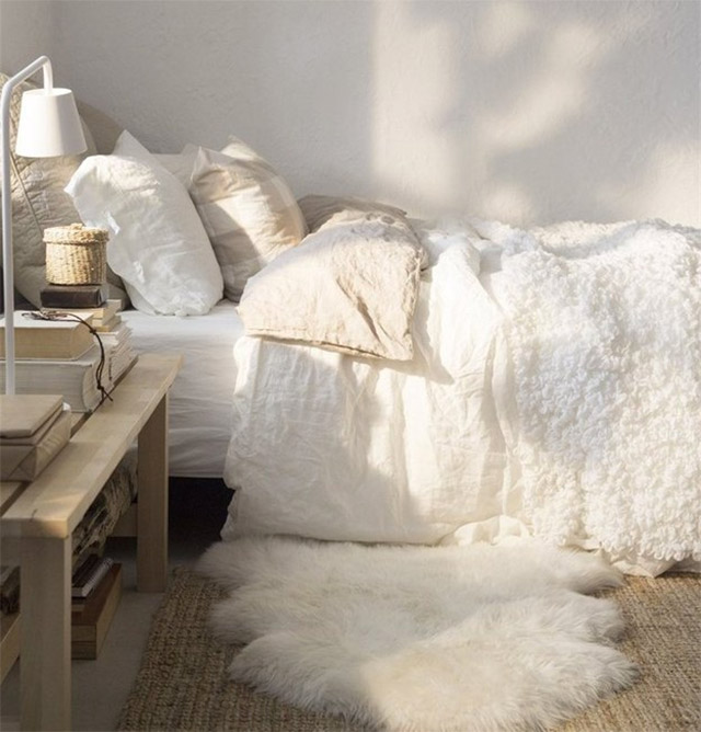 Dormitorio con cama en tonos blancos y crudos. Sábanas blancas con almohadas y cojines en blanco y beige muy claros, piecera blanca tejida en buclés. Bajada de cama de piela blanca sobre alfombra de yute.