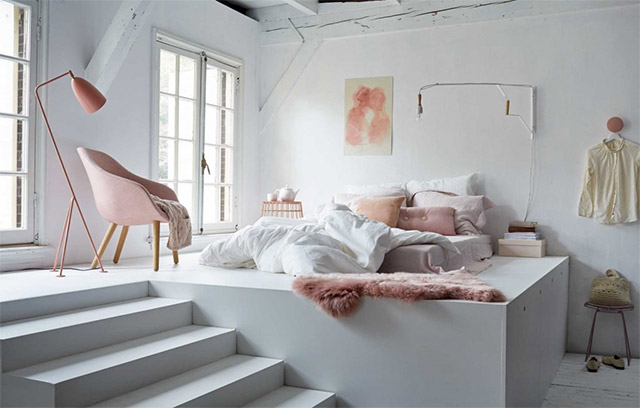 Dormitorio en blanco y rosa. La habitación es completamente blanca de paredes a techo y piso. En una plataforma más elevada, a la que se accede por una escalera, se encuentra la cama, al piso, con cubre colchón en rosa cuarzo, cobertor blanco, cojines y almohadones blancos y en tonos rosa. A un costado y cerca de los ventanales, una lámpara de pie en color rosa, una poltrona de tela en rosa cuarzo y patas de madera y una mesita auxiliar rosa y sobre la mesa un juego de tetera y tacita blancas.