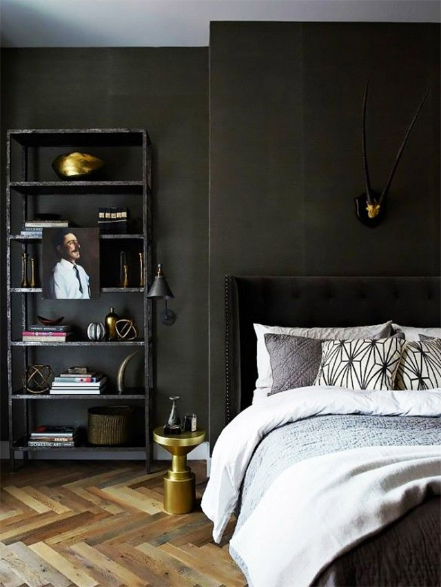 Dormitorio en tonos negros. Paredes negras, piso de parquet, cielo blanco. Mueble estante negro a un costado de la cama con accesorios dorados, como fuentes y candelabros. Cama de respaldo de tela negra con remaches metálicos, ropa de cama en grises muy claros, cojines blancos, negro y gris. Una pequeña mesita velador dorada.