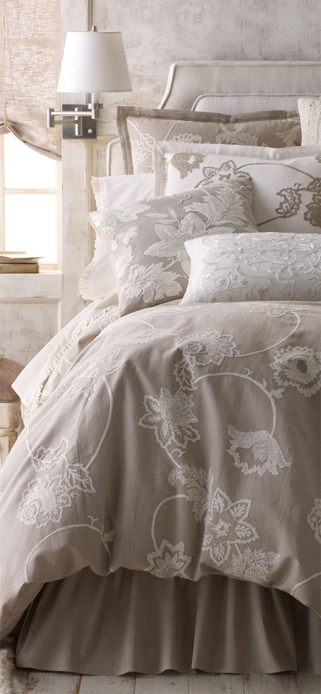 Cama con textil en beige claro y blanco con bordados de flores en los mismos colores. A un costado de la cama y desde el muro, una pequeña lámpara de cuerpo metálico flexible y pantalla blanca.