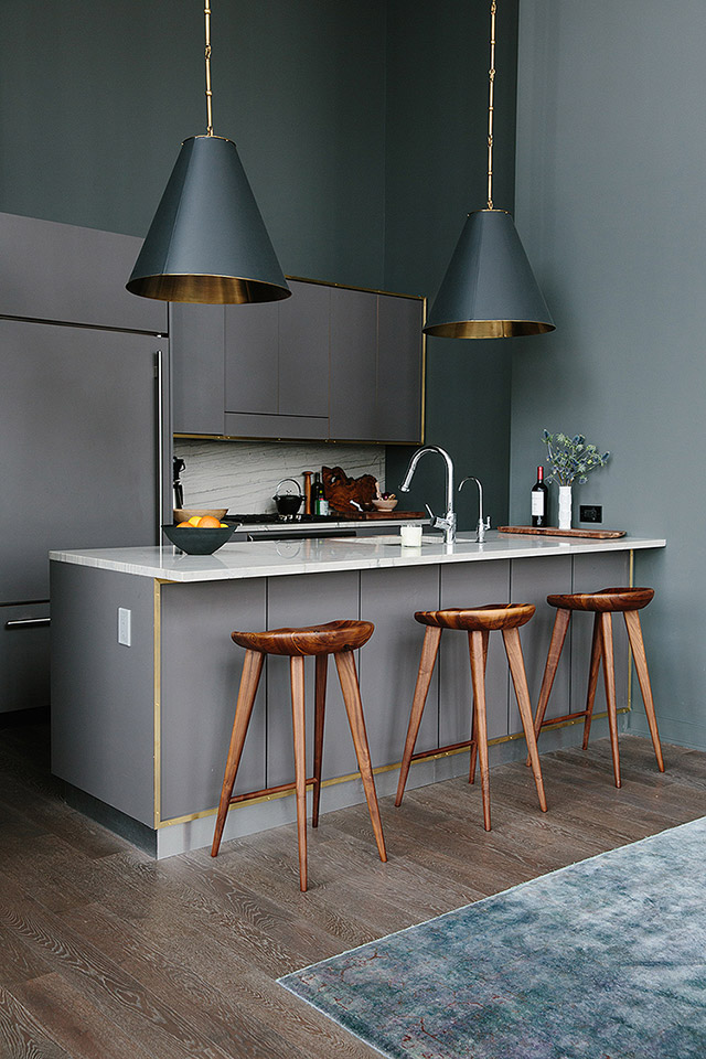 Cocina americana en tonos grises en los muros, piso de madera, alfombra gris con matices rojos. Pisos altos de madera y sobre el mesón de la cocina dos lámparas de pantalla negra e interior cobre.