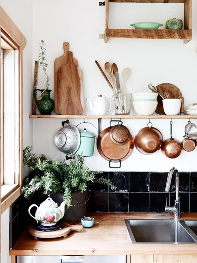 Cocina con repisa de madera sobre al lavaplatos. De la repisa cuelgan ollas y sartenes de cobre. Sobre la repisa pocillos y fuentes blancas y utensilios de cocina de madera, como tablas de picar y cucharas.