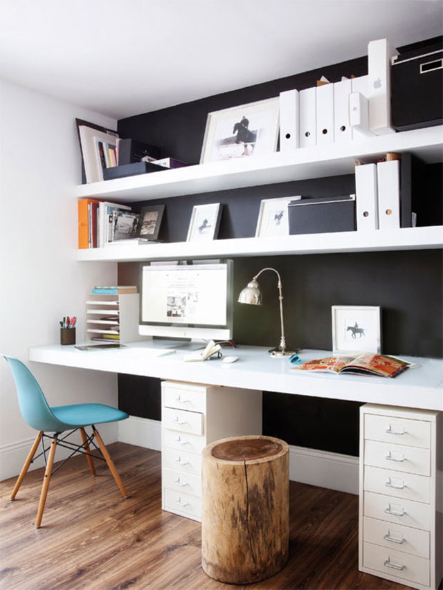 Escritorio completamente incorporado al muro, con una mesa y cajoneras en blanco y repisas blancas sobre muro negro. La silla es una Eames de respaldo calipso y patas de madera, a un costado un trozo de tronco que sirve de piso.