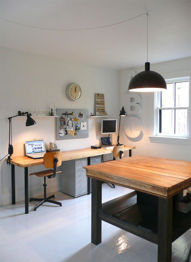 Taller con mesón escritorio con cubierta de madera gruesa natural para dos puestos de trabajo. Una mesa cuadrada con cubierta de madera natural al centro del taller. La decoración es mínima con un par de repisas, un mural y un reloj.