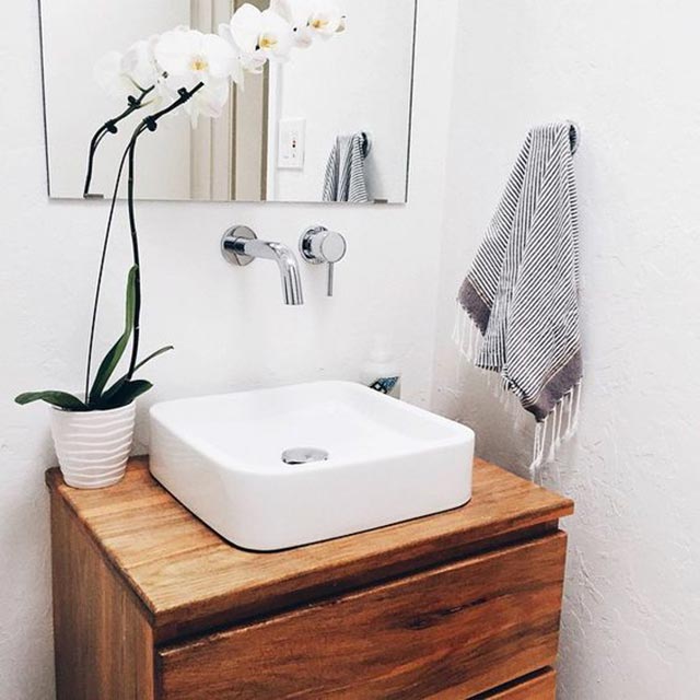 Baño con el mueble vanitorio de madera, en la cubierta al costado del lavamanos decora una maceta con orquídeas.