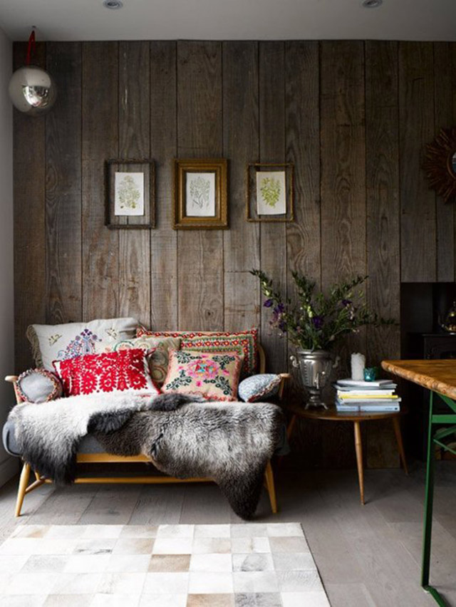 Habitación con pared de listones gruesos de madera oscura puestos de manera vertical, sillón de estructura de madera, mesa lateral redonda de tres patas de madera El sofá decorado con pieles grises y cojines bordados. La pared decorada con tres ilustraciones de flores enmarcadas.