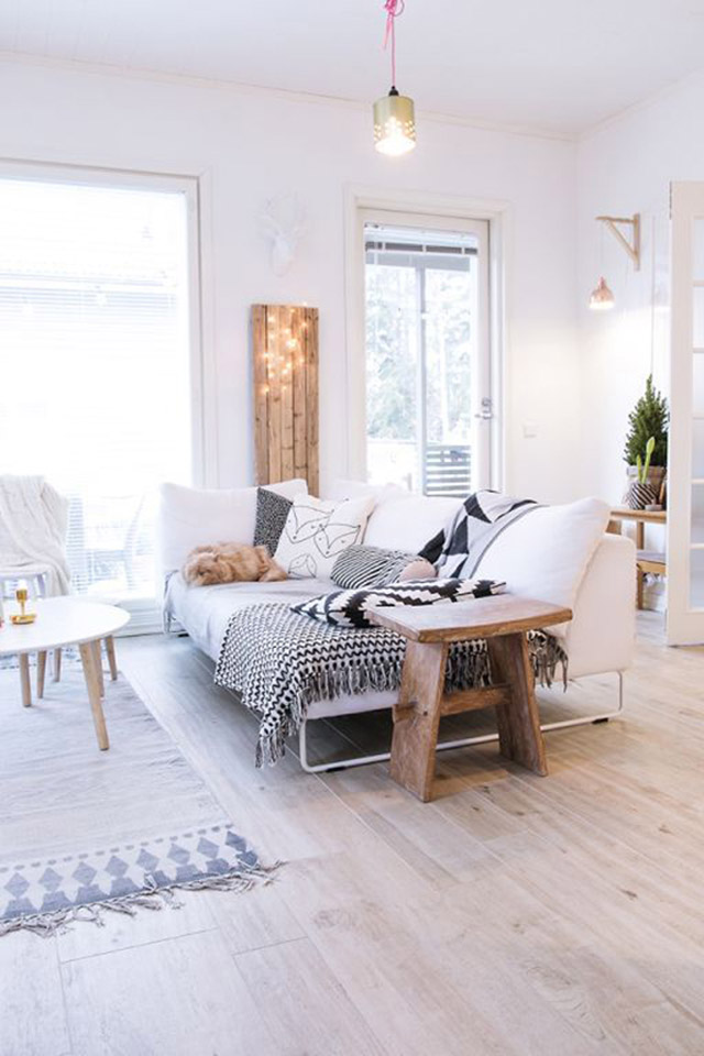 Sala de estar estilo nórdico con piso de madera, mesa de centro redonda con patas de madera, banqueta estilo rústico de madera al costado del sofá blanco. Sobre el muro, entre dos ventanales, un panel de madera decorado con una guirnalda de luces.