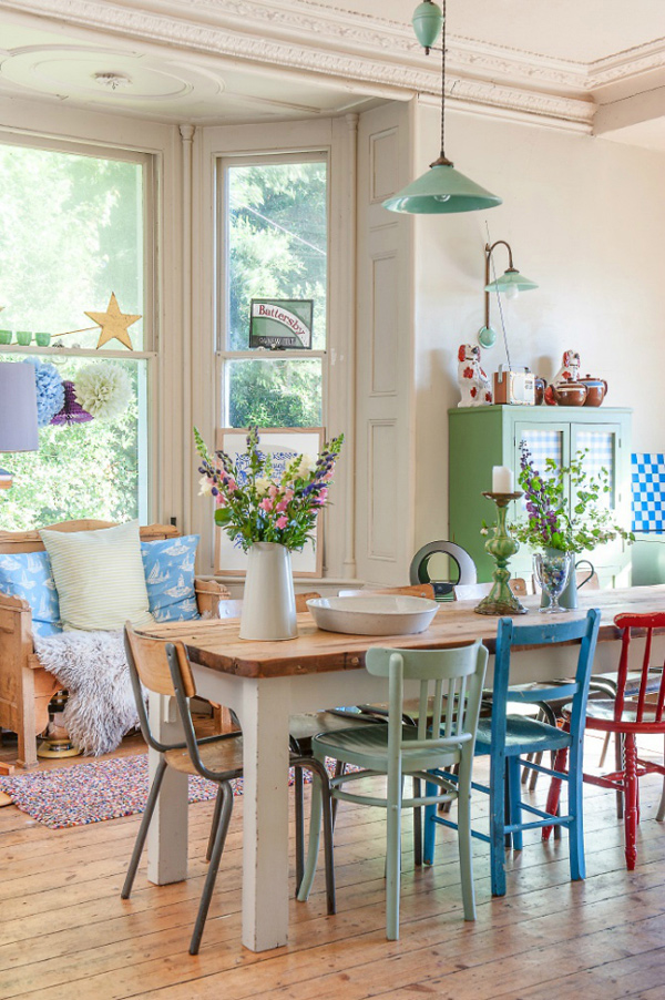 Comedor muy colorido con mesa de madera de patas gruesas blancas y cubierta de madera color natural. Las sillas son de madera y colores verde claro, azul y roja, para complementar con sillas de estructura metálica y asiento y respaldo de madera barnizada.