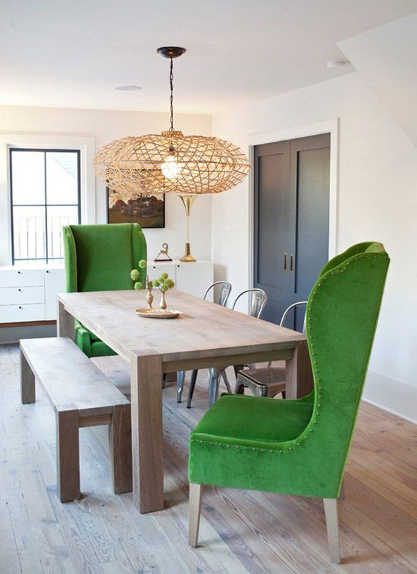 Comedor con mesa de madera color natural de líneas simples con banqueta del mismo estilo en uno de los costados, tres sillas Tolix metálicas en frente y dos sillones de tela verde y patas de madera en las cabeceras. Lámpara de fibra natural de forma circular colgando sobre la mesa.