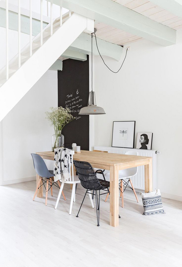 Comedor estilo nórdico con mesa de madera clara y líneas simples, cinco sillas, un par de sillas Eames en blanco y celeste; sillas Lilla Aland, una blanca y la otra en madera natural; y una variante de la silla Eames con el respaldo y asiento tejido en negro. Del techo cuelga una lámpara de estilo industrial.