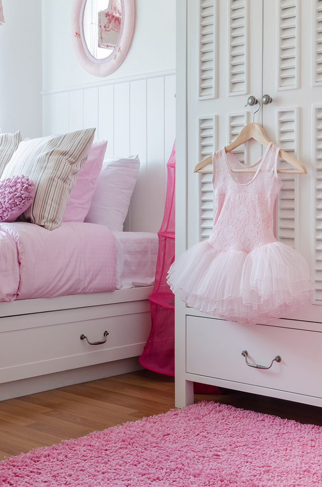 Dormitorio de niña. Cama con cajonera y ropa de cama en color rosa y blanco, al costado un clóset y de las manillas de las puertas del clóset un colgador con un tutú rosado.