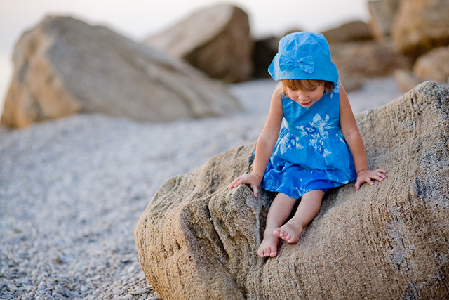 Niña sentada en una roca a la orilla de la playa con vestido y gorro azul.