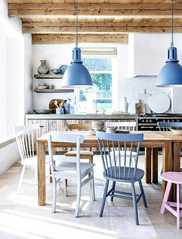 Comedor y cocina de techo con vigas a la vista, muros blancos. Mesa de comedor de madera natural y sillas blancas y tonos de azul serenity. También podemos ver un piso en rosa. Las lámparas del techo, de tipo industrial, de pantallas metálicas y color azul.