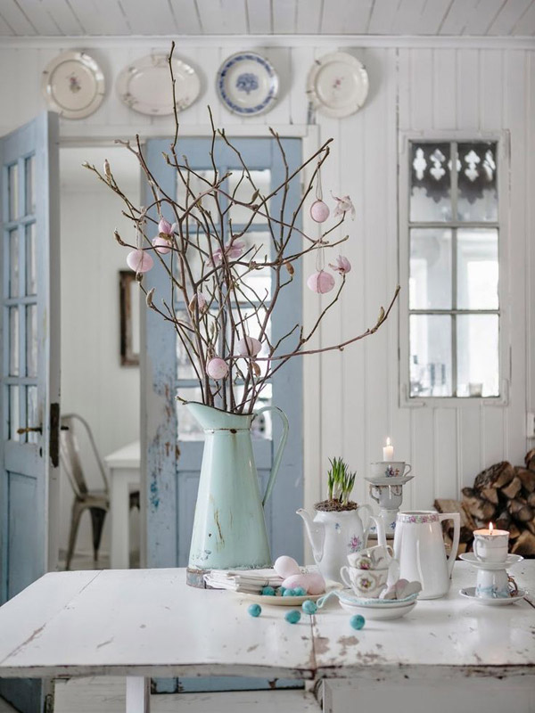 Mesa de comedor blanca de madera, muy shabby chic, sobre la mesa la loza es blanca con detalles en celeste y rosa, algunas de las tazas utilizadas como candelabros. El jarrón de latón con ramas es de color celeste. Al fondo, separando el comedor de la siguiente habitación, unas puertas de color serenity.